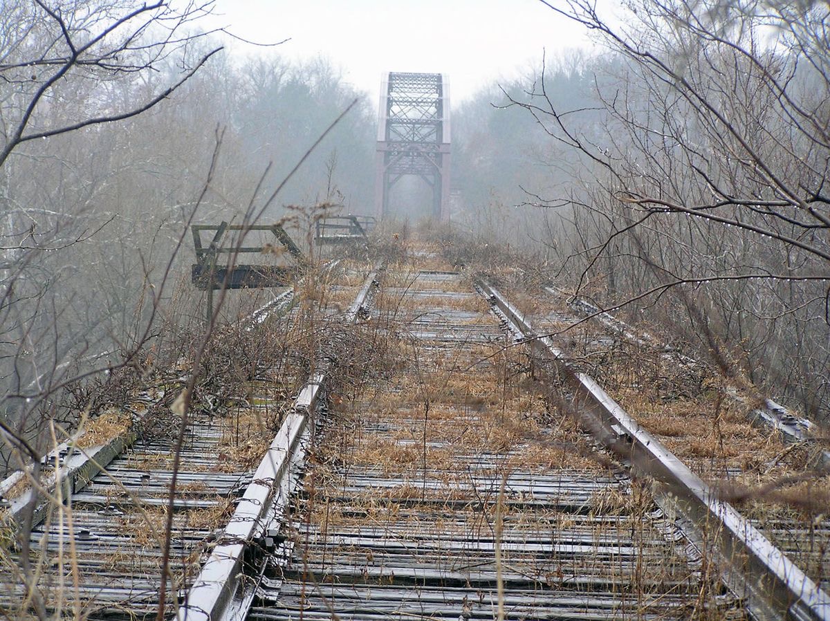 An abandoned railway trestle, rusted rails disappearing into fog toward a steel truss bridge, wooden ties overgrown with brush and dried grass.